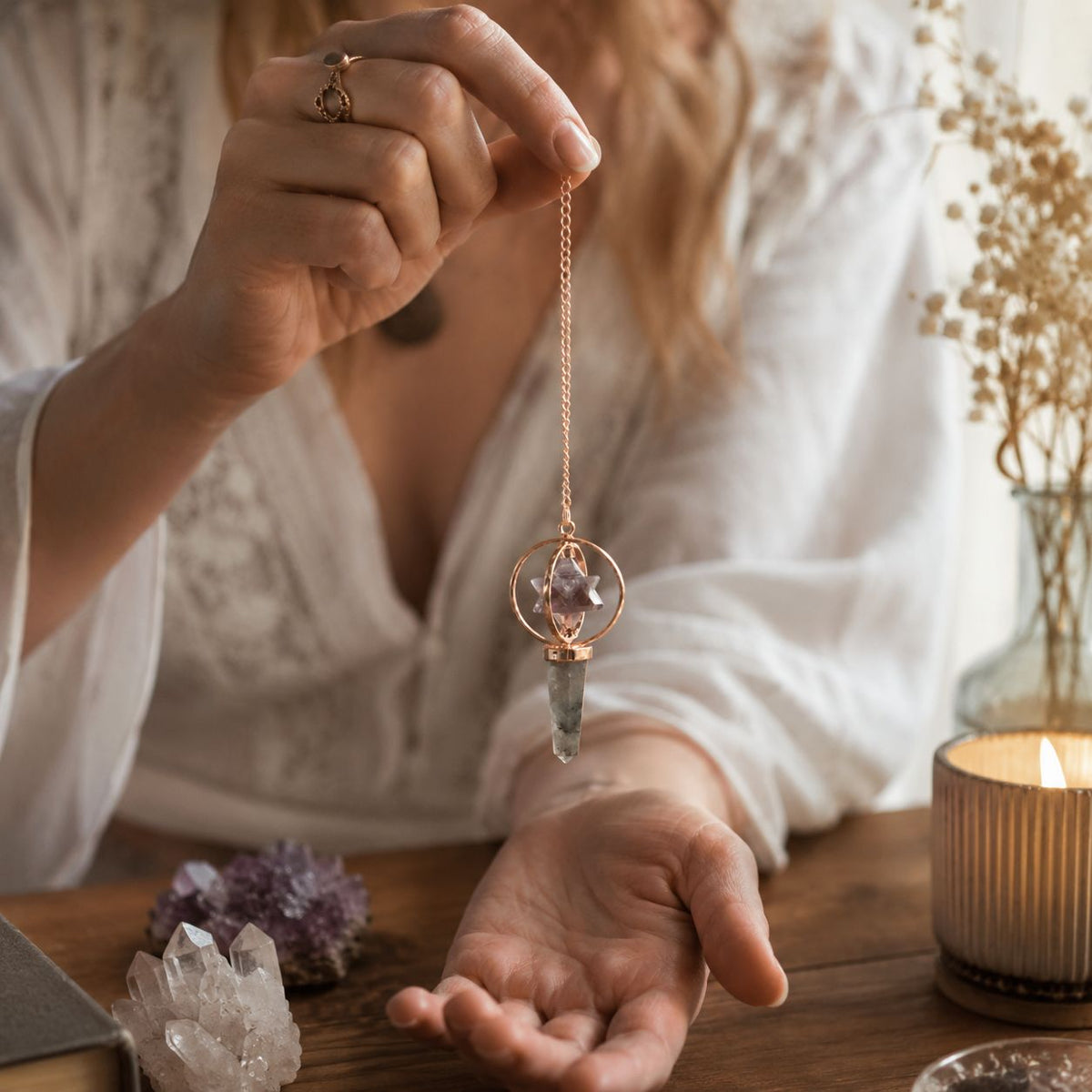 Person holding a crystal pendant over their palm with a candle and crystals on a table.