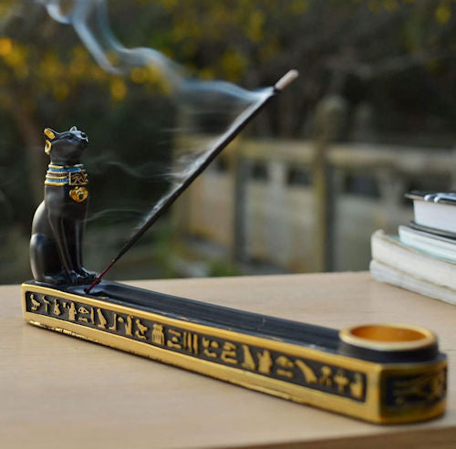 An Egyptian-style black cat incense holder with gold hieroglyphic details, featuring a smoking incense stick resting on its back, set on a wooden table with a blurred outdoor background.