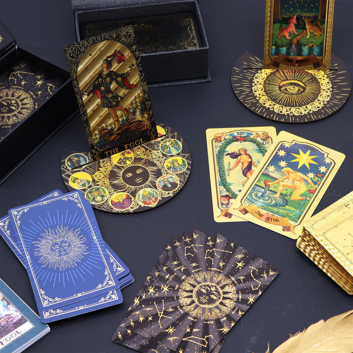A tarot card deck set arranged on a dark surface, including multiple illustrated cards, several black boxes, a gold-toned stand, a circular decorative base, and blue-backed cards. The cards feature fantasy and celestial artwork with visible titles such as Fool, The Fool, The Star, and The Queen.