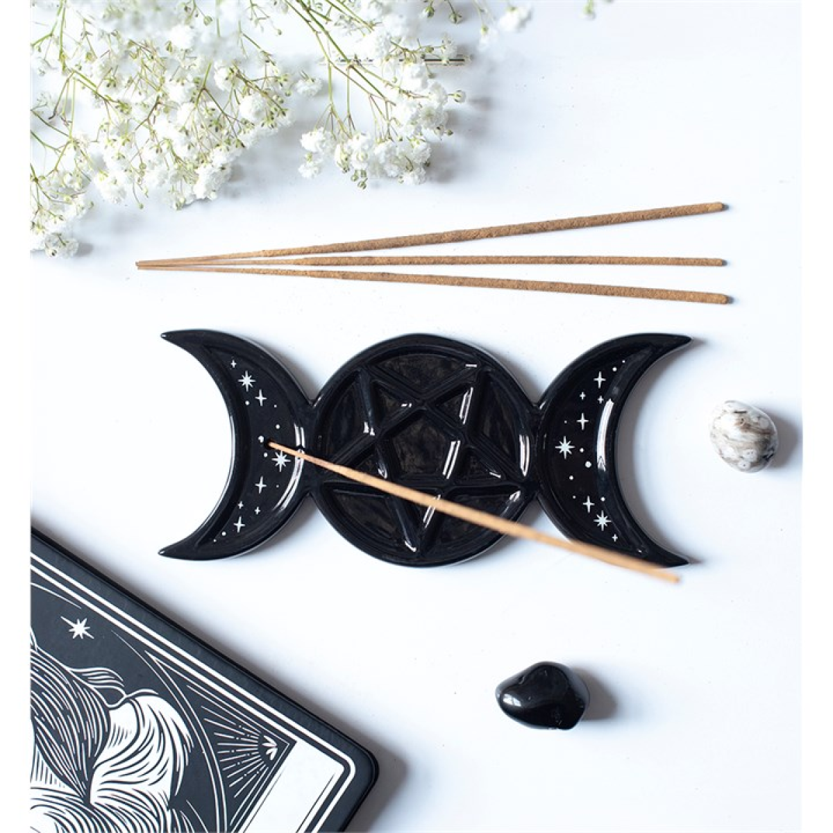 A black triple moon and pentagram altar piece with three crescent moons and a central pentagram, styled on a white background with incense sticks, baby's breath flowers, a tarot card corner, and small stones.