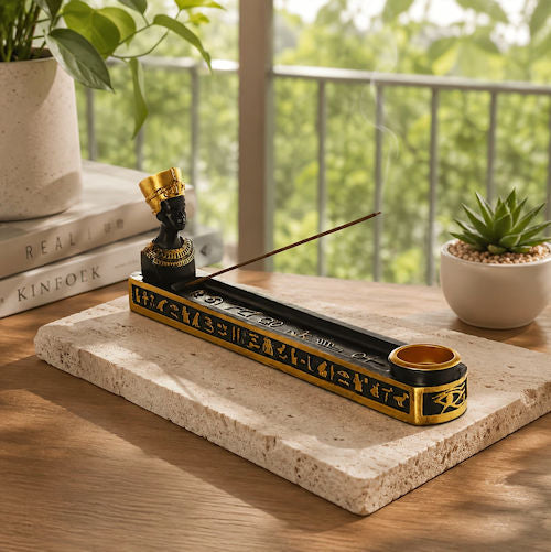 A black and gold incense holder with a figurine and incense stick, displayed on a stone tray on a wooden table with a blurred outdoor garden background.